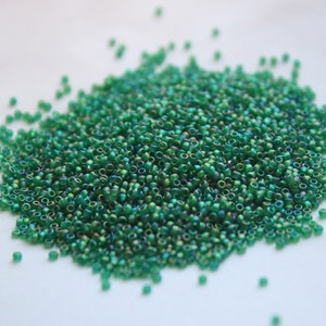 May include: A large pile of small, round, green iridescent beads. The beads are scattered on a white surface.