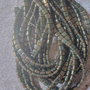 May include: A pile of dark green iridescent glass beads, each bead is square shaped with rounded edges.