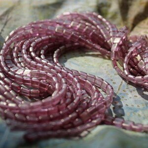 May include: A strand of iridescent pink glass beads, each bead is a square shape with rounded edges.