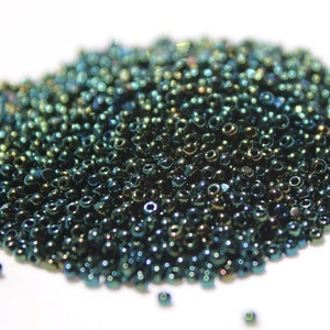 May include: A pile of small, iridescent teal seed beads. The beads have a metallic sheen and are scattered on a white surface. These beads are commonly used for jewelry making and crafting projects.