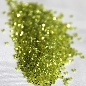 May include: A large pile of small, light green glass seed beads. The beads are iridescent and have a metallic sheen.