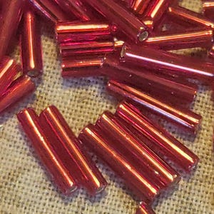 May include: A pile of red glass beads, some are long and thin, others are shorter and thicker. The beads are shiny and have a metallic finish.