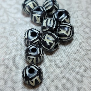 May include: A collection of black round beads with cream-colored geometric patterns. Each bead has a hole through the center. The beads are scattered on a light gray patterned surface. These beads are suitable for jewelry making and crafts.