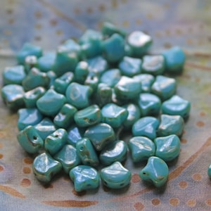 May include: A pile of turquoise blue glass beads in the shape of a teardrop. The beads have a slightly iridescent finish.