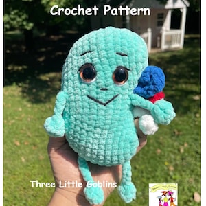 May include: A crocheted teal-coloured monster with large brown eyes and a small black smile. The monster is holding a small crocheted blue and white snowman. The image includes the text "Three Little Goblins" and a logo for "Three Little Goblins".