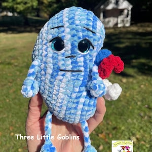 May include: Blue and white crocheted plush toy with a sad expression and large blue eyes. The toy has two arms and two legs, and is holding a red, white, and blue crocheted ball. The toy is labeled "Three Little Goblins".