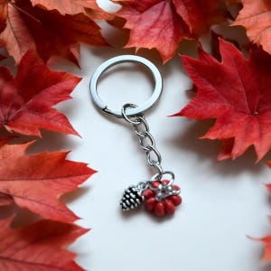 May include: A silver keychain with a red pumpkin and a pine cone charm. The keychain is surrounded by red maple leaves.