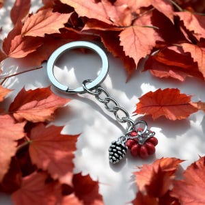 May include: A silver keychain with a red and silver charm. The charm is shaped like a pine cone and a small pumpkin. The keychain is surrounded by red autumn leaves.