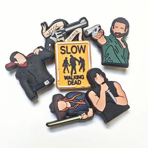 May include: A selection of brightly coloured shoe charms depicting characters and symbols from "The Walking Dead." The charms include images of Rick Grimes with a gun, a sign that says "SLOW WALKING DEAD," and other characters. These charms are designed to be attached to shoes.