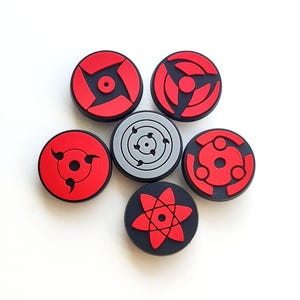 May include: A set of six circular shoe charms featuring various red and black graphic patterns. Five charms have red designs on black backgrounds, while one central charm features a grey background with black line art details.