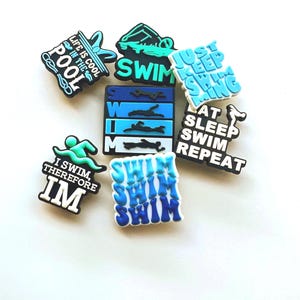May include: A selection of vibrant shoe charms with swimming-related designs. The charms display phrases such as "Swim," "Life is Cool in the Pool," and "I Swim Therefore I'm." The colour scheme includes blue, white, and green.
