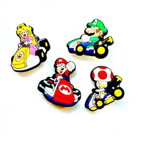 May include: Four colourful cartoon character charms in go-karts. Characters include Princess Peach in a yellow kart, Luigi in a green kart, Mario in a red kart, and Toad in a red and white kart. The charms are on a white background.
