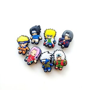 May include: A collection of seven small, colourful rubber shoe charms featuring stylised cartoon characters from the Naruto series. Each charm depicts a different character with distinct hair colours, outfits, and headbands, rendered in a flat, 2D design.