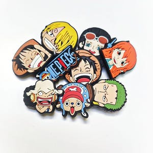 May include: A selection of colourful cartoon character charms, depicting various faces with distinct hairstyles and expressions. The word "ONE PIECE" is visible on a blue and white charm. The charms are arranged on a white surface.