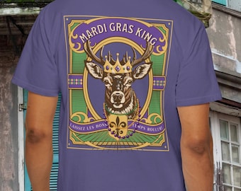 May include: A purple t-shirt featuring a detailed graphic of a deer wearing a crown, with the words "Mardi Gras King" above and French text below. The design includes green, gold, and purple accents, and a fleur-de-lis symbol.