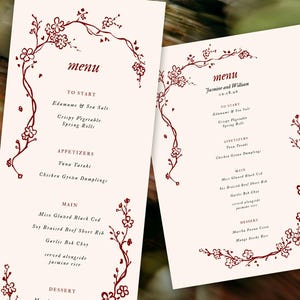 Op de afbeelding: Twee crèmekleurige trouwmenu's met bordeauxrode bloemenaccenten. De menu's tonen een lijst met gerechten, waaronder voorgerechten en hoofdgerechten. Het woord "menu" staat bovenaan en de namen "Jasmine en William" zijn ook zichtbaar.