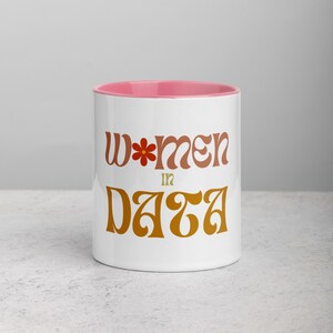 Puede incluir: Taza de cerámica blanca con borde rosa. La taza tiene un diseño gráfico que dice "Women in Data" en una fuente retro con una flor en la "W".