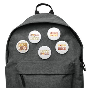 Könnte beinhalten: Ein grauer Rucksack mit fünf runden Knöpfen, die an der Vorderseite befestigt sind. Die Knöpfe haben eine Schriftart im Retro-Stil und sagen "Data Diva" und "Women in Data".