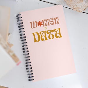 Może przedstawiać: Różowy notes w spirali z tekstem "Women in Data" w retro czcionce z kwiatem w "W".