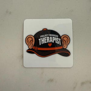 May include: A square sticker featuring a black baseball cap with an orange trim and two large ears protruding from the sides. The text PROJECT MANAGER THERAPIST is printed in white on the cap, with a small orange heart below the word THERAPIST.
