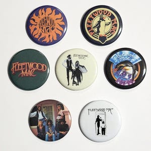 May include: A collection of seven round Fleetwood Mac band buttons. The buttons feature various album art and logos, including a sunburst design, a penguin, and band member portraits. Colors include orange, green, black, and white.