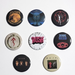 May include: A collection of seven round pinback buttons featuring various designs and the band name "RUSH." The buttons display album art and band imagery, including an owl, a figure within a star, and band member portraits. Colors include black, white, red, and gold.