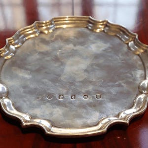 Pode incluir: Uma bandeja oval prateada com uma borda decorativa ondulada. A bandeja tem uma superfície reflexiva e parece ser feita de metal. Pequenas marcas são visíveis perto do centro. Provavelmente usado para servir ou exibir.