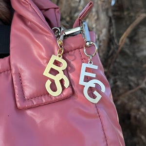 Può includere: Una borsa imbottita color rosa polvere con due ciondoli in metallo fissati alla cerniera. Uno è un monogramma color oro con le lettere B e S, l'altro è un monogramma color argento con le lettere E e G, entrambi appesi a piccoli ganci.