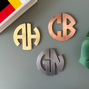 Może przedstawiać: Trzy metalowe podstawki z monogramem w kolorze złotym, miedzianym i srebrnym. Każda podstawka ma inny dwuliterowy monogram w okrągłym kształcie. Podstawki są prezentowane na szarej powierzchni, z książką i rośliną w tle.
