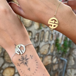 Op de afbeelding: Twee armbanden met ronde hangers. Eén is zilverkleurig met de initialen "MC" uitgesneden, en de andere is goudkleurig met een gestileerd ontwerp. Beide armbanden zitten aan fijne kettingen.