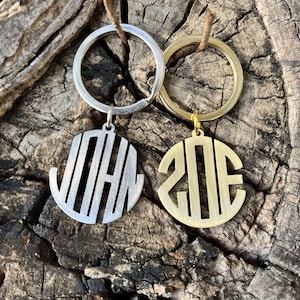 May include: Two personalised keyrings with circular pendants. One is silver with the name "JOHN" cut out, and the other is gold-toned with the initials "ZDE". Both have key rings and are displayed on a textured wooden surface.