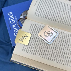 Può includere: Due segnalibri quadrati in metallo appoggiati su un libro aperto. Uno è color oro con le lettere RTV ritagliate, l'altro è color argento con le lettere GS ritagliate. Entrambi presentano angoli arrotondati e un design geometrico piatto.