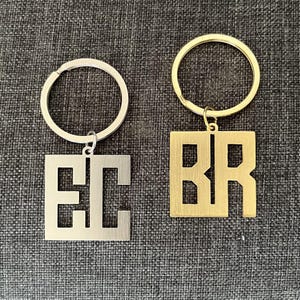 May include: Two metal keyrings with square pendants. One is silver with the letters "EC", the other is gold with the letters "BR". Both keyrings have a circular ring attached to the top of the pendant.