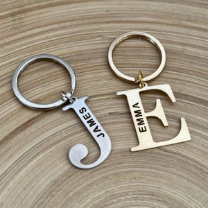 Pode incluir: Dois chaveiros personalizados. Um é prateado, em forma da letra "J" com "JAMES" gravado. O outro é dourado, em forma da letra "E" com "EMMA" gravado. Ambos têm argolas para chaves.