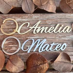 Może przedstawiać: Dwa breloczki do kluczy z imionami "Amelia" w kolorze złotym i "Mateo" w kolorze srebrnym, umieszczone na drewnianej powierzchni. Każde imię jest w ozdobnej czcionce i przymocowane do metalowego kółka. Tło zawiera suche liście.
