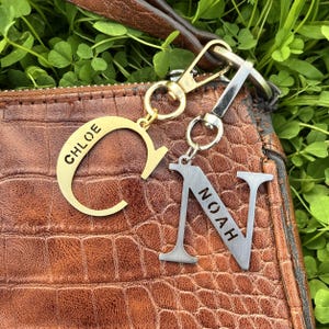 Könnte beinhalten: Zwei personalisierte Schlüsselanhänger mit Namen. Einer ist ein goldfarbener Buchstabe "C" mit dem eingravierten Namen "CHLOE". Der andere ist ein silberfarbener Buchstabe "N" mit dem eingravierten Namen "NOAH". Beide Schlüsselanhänger haben einen Verschluss.
