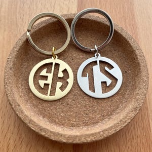 May include: Two personalized keychains with initials. One keychain is gold-colored with the initials "EB" and the other is silver-colored with the initials "TS". Both keychains are round and sit on a brown, circular surface.