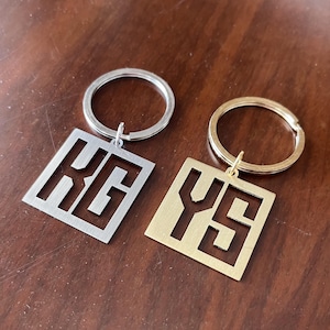 May include: Two keyrings with square pendants. One is silver with the letters "KG" cut out, and the other is gold with "YS". Both have a circular key ring attached.