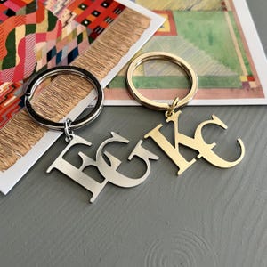 Peut inclure: Deux porte-clés avec des pendentifs en forme de lettres. L'un est argenté avec les lettres "E G C" et l'autre est doré avec les lettres "K C". Les porte-clés sont sur une surface grise avec des œuvres d'art colorées en arrière-plan.