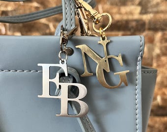 Op de afbeelding: Close-up van een lichtblauwe handtas met twee metalen bedels. Eén bedel is zilverkleurig en heeft de letters "FB", de andere is goudkleurig met de letters "NG". De tas heeft een handvat aan de bovenkant en een schouderriem.