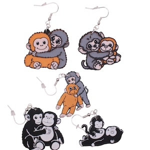 May include: A collection of five dangle earrings featuring illustrated pairs of monkeys in various poses. The designs include orange and grey monkeys hugging, as well as black and white monkeys in different positions, all attached to silver-tone hooks.