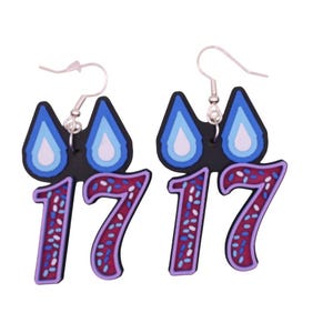 May include: A pair of dangle earrings featuring a black, blue, and white design. The earrings have a raindrop shape at the top and the number "17" in a burgundy color with white and blue speckles. The earrings hang from silver hooks.