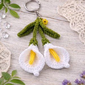 Peut inclure: Un porte-clés fait à la main avec deux fleurs de calla lily blanches et des centres jaunes, avec des tiges vertes. Le porte-clés a un anneau argenté. Fond en bois clair.