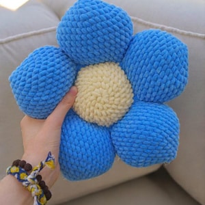 Peut inclure: Un coussin fait à la main en forme de fleur, bleu et jaune. Le coussin a six pétales bleus entourant un centre jaune. La texture semble douce et moelleuse. Une main tient le coussin.