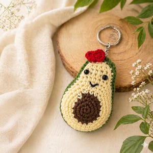 Peut inclure: Porte-clés avocat crocheté à la main. L'avocat a un extérieur vert, un intérieur jaune clair et un noyau marron. Il présente un visage souriant et un accent rouge. Le porte-clés est en argent.