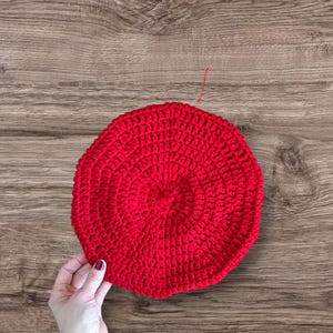 Peut inclure: Un béret rouge vif, fait à la main au crochet. Le béret est rond avec un motif texturé et circulaire. Le chapeau est tenu par une main sur un fond en bois.