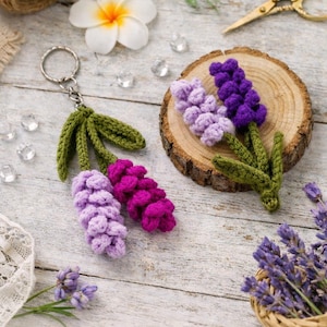 Peut inclure: Des porte-clés et des fleurs décoratives en crochet faits à la main, représentant des fleurs de lavande violettes et vertes. Les porte-clés présentent un motif de fleur de lavande avec des feuilles vertes. Les fleurs décoratives sont exposées sur une tranche de bois.