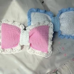 Peut inclure: Trois coussins en forme de nœud papillon. Deux sont roses et blancs, et un est bleu et blanc. Les coussins ont un aspect matelassé texturé et sont bordés d'une bordure en crochet à volants. Ils sont disposés sur une surface blanche.