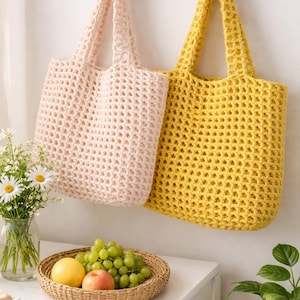 Peut inclure: Deux sacs fourre-tout en crochet faits à la main, un rose clair et un jaune, sont suspendus à un crochet mural en osier. Les sacs ont un motif de tissage ouvert. Un panier de fruits et un vase de fleurs sont sur une table.