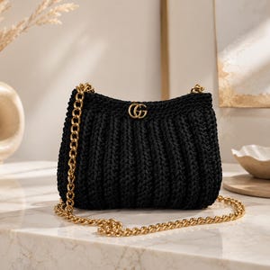 Peut inclure: Un sac à main noir en crochet présentant une texture côtelée et une bandoulière en chaîne métallique dorée. L'avant du sac est orné d'un petit emblème logo GG en métal doré centré, offrant un contraste marqué avec le tissu sombre.
